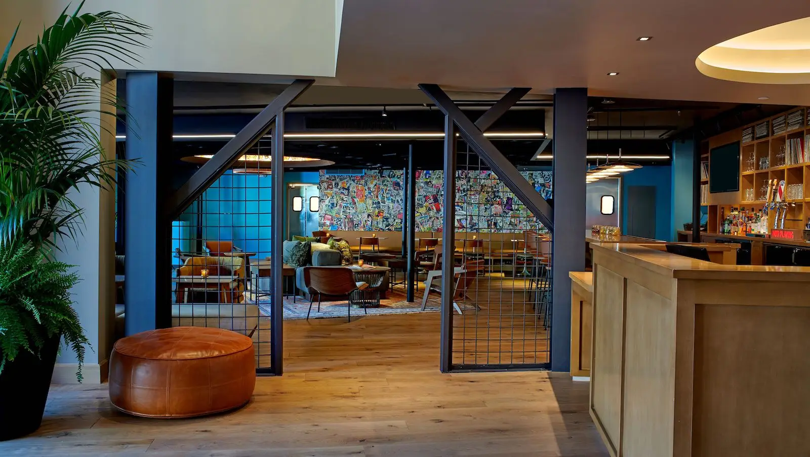 A modern, open-plan lounge area at Hotel Ziggy boasts wooden flooring, a colorful mural wall, and stylish furniture. It includes leather and fabric chairs, a round cushioned stool, and tables. The space features ceiling lights, decorative plants, and a partial view of a bar area on the right.
