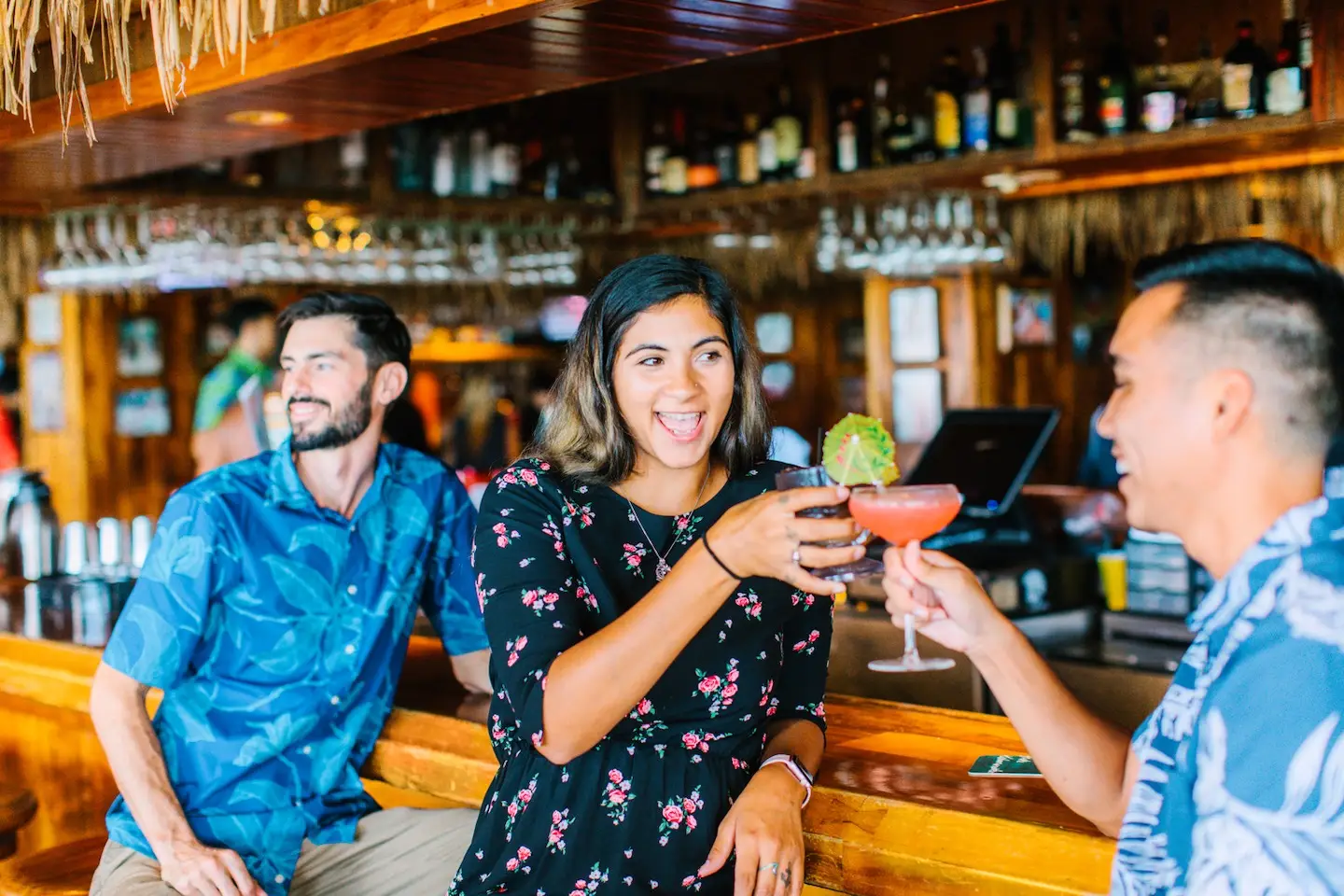 Three people at a bar, with two of them clinking cocktails and smiling. The person on the left is seated and laughing, while the other two, standing, are holding brightly colored drinks. The relaxed vibe resembles an Outrigger music venue or a Hawaii home, with bottles and glasses on display in the tropical ambiance.