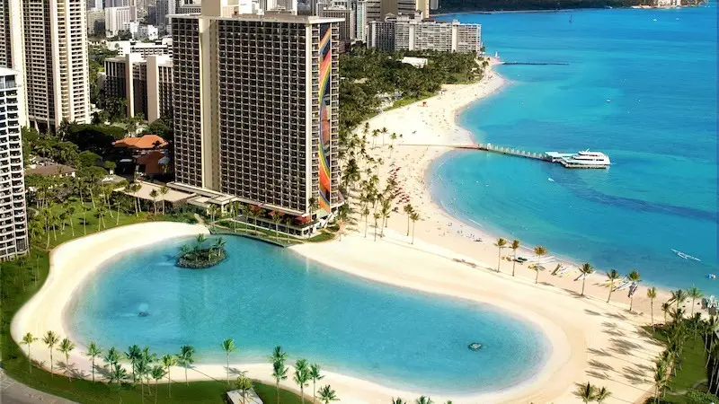 A high-rise hotel with a colorful design overlooks a scenic, tropical beach with white sand and turquoise water. A large lagoon with a small island is in the foreground, surrounded by palm trees. In the background, other hotels and buildings line the shoreline, making it one of the best hotels with pools in Hawaii.
