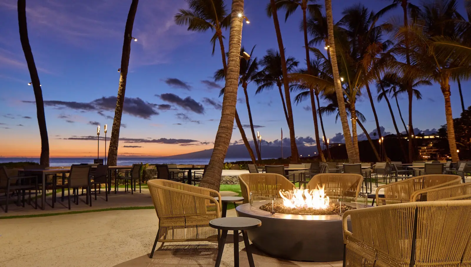 Outdoor beachside dining area during sunset at Kā‘anapali Beach Hotel, with a central fire pit surrounded by wicker chairs and tables. Palm trees are silhouetted against the colorful Hawaiian sky, and the ocean is visible in the background. The atmosphere is serene and inviting.