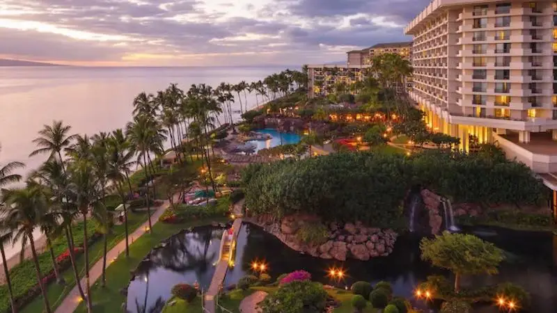 A luxurious beachfront resort at sunset perfect for a family getaway. The scene features a high-rise hotel, palm trees, tropical gardens, illuminated pathways, a pool area, and a serene pond with a waterfall. The ocean and Hawaii’s colorful twilight sky are visible in the background.