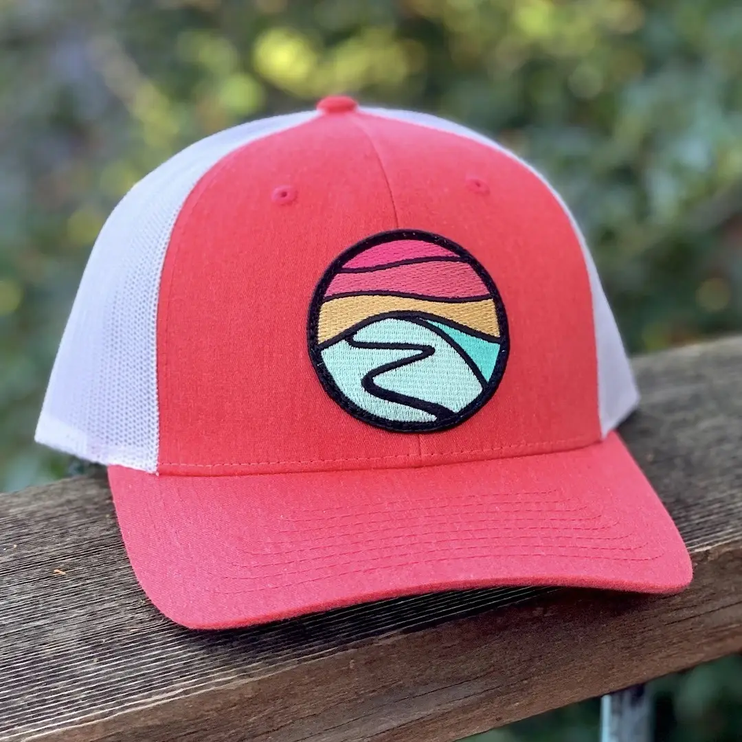 A coral-red trucker hat with a white mesh back is displayed on a wooden ledge. This piece of apparel features a circular patch on the front, showcasing a stylized landscape design in shades of pink, yellow, and blue, reminiscent of a Tommy Breeze sunset over water. Green foliage is blurred in the background.