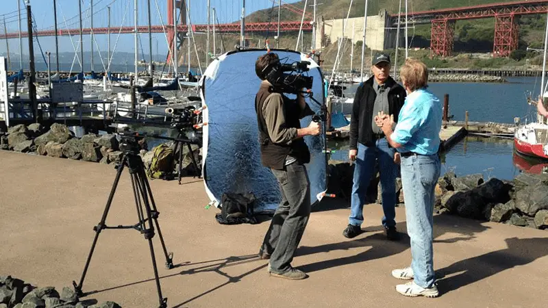 Three people are filming a scene on a dock with boats and water in the background. The setup includes a camera on a tripod, a man holding another camera, and a large reflector. Bay Area Backroads' Doug McConnell is present, with the Golden Gate Bridge visible in the distance.