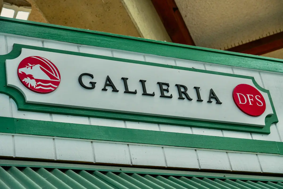 A sign for "Galleria DFS" is mounted on a green-trimmed building. The sign has black text on a white background, with decorative green border details. Logos featuring an airplane and a palm tree in red circles are on the far left and right sides of the sign, offering tips on Duty Free Shopping in Maui airport Hawaii.