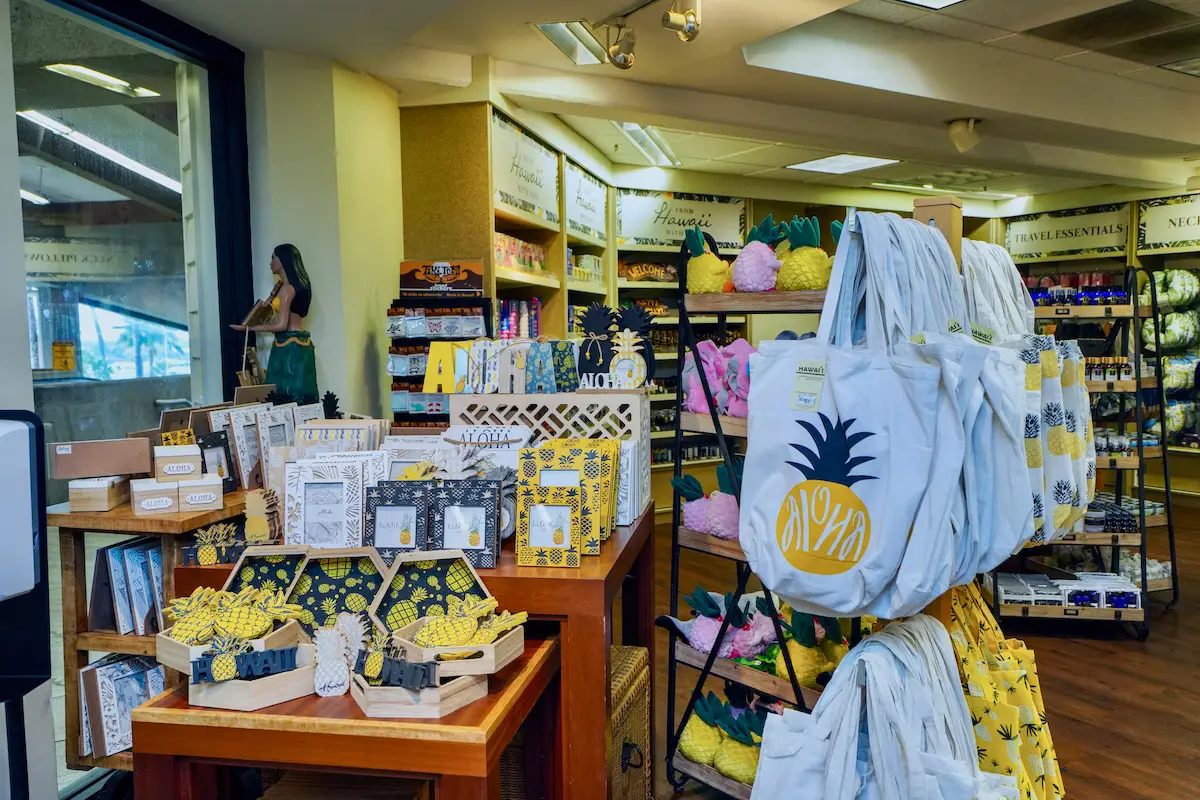 A brightly lit store featuring Hawaiian-themed merchandise and tips on Duty Free Shopping in Maui airport Hawaii. Tote bags with pineapple designs hang prominently, while various items like postcards, snacks, and other souvenirs are displayed on wooden tables and shelves. A statue of a hula dancer is seen in the background.
