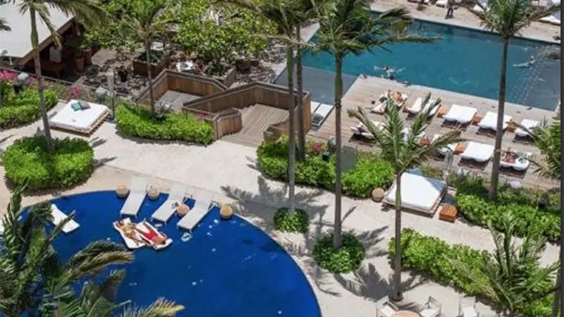 Aerial view of a tropical resort featuring a large, curved blue pool surrounded by palm trees, lounge chairs, and lush greenery. Guests are seen relaxing by the pool. A wooden deck and a second rectangular pool are visible in the background, making it one of the best hotels on Oahu.