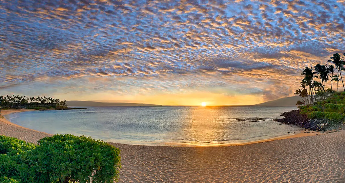 A serene beach scene at sunset near The Napili Kai Beach Resort. The sky features a mix of vibrant colors and scattered clouds. The sun is setting near the horizon, reflecting golden light on the calm ocean. Lush greenery lines the sandy shore, and palm trees dot the landscape on the right, evoking a true Hawaii home feel.