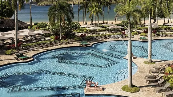A luxurious outdoor swimming pool is surrounded by lounge chairs and tall palm trees. Beyond the pool area, a sandy beach and blue ocean are visible. The scene is drenched in sunlight, creating an inviting atmosphere for relaxation, epitomizing the allure of the best hotels with pools Hawaii offers.