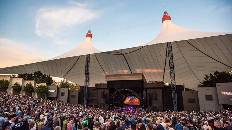 A large outdoor concert venue with a white tent-like roof structure. The stage is set up under the canopy, and a large audience fills the seating area and lawn. The sky is clear with a few clouds, indicating an evening event. Trees are visible around the venue; one of the best things to do in the South Bay this March.