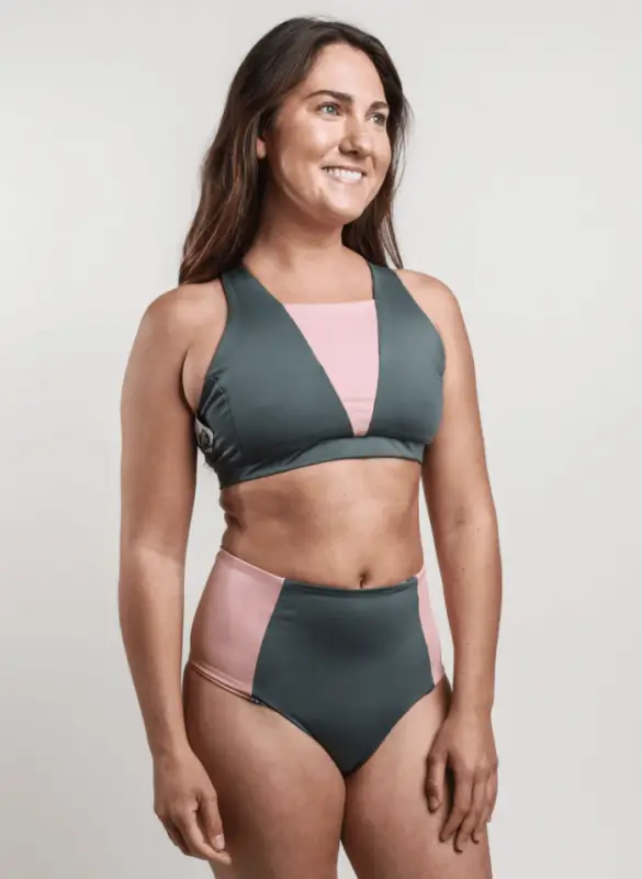 A person with long dark hair is wearing a two-piece swimsuit. The dark green and pink swimsuit, featuring a high-waisted bottom and a top with cut-out details, embodies the spirit of Bikinis That Show You Care About Planet. The person is smiling and facing slightly to the right against a plain background.