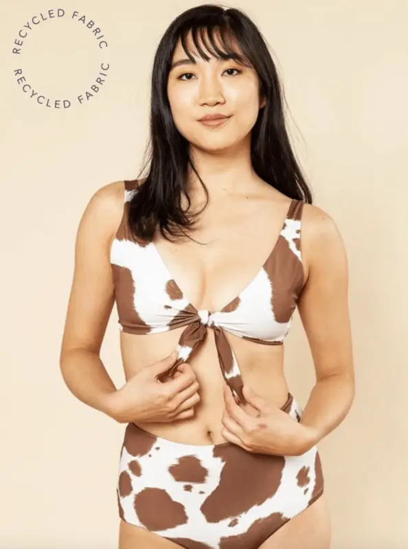 A woman with long black hair stands confidently in a cow print bikini against a plain background. The bikini top ties in front, showcasing the design from Bikinis That Show You Care About Planet. The text “Recycled Fabric” appears twice in a circular design on the left side of the image.