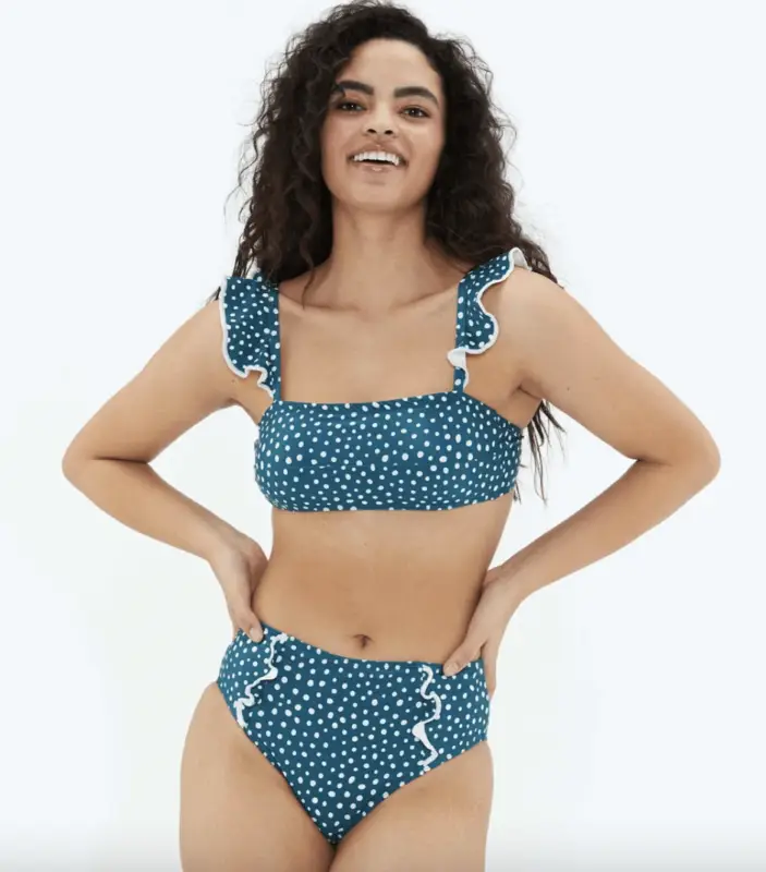 A woman with long wavy hair is wearing a blue high-waisted bikini with white polka dots and ruffled straps, standing with her hands on her hips, smiling against a plain white background. Embrace bikinis that show you care about the planet.