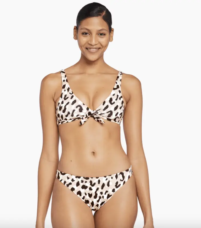 A woman is modeling a leopard print bikini with a knot detail on the top. She has her hair slicked back and is smiling at the camera against a plain white background, effortlessly showcasing one of the "Bikinis That Show You Care About Planet".