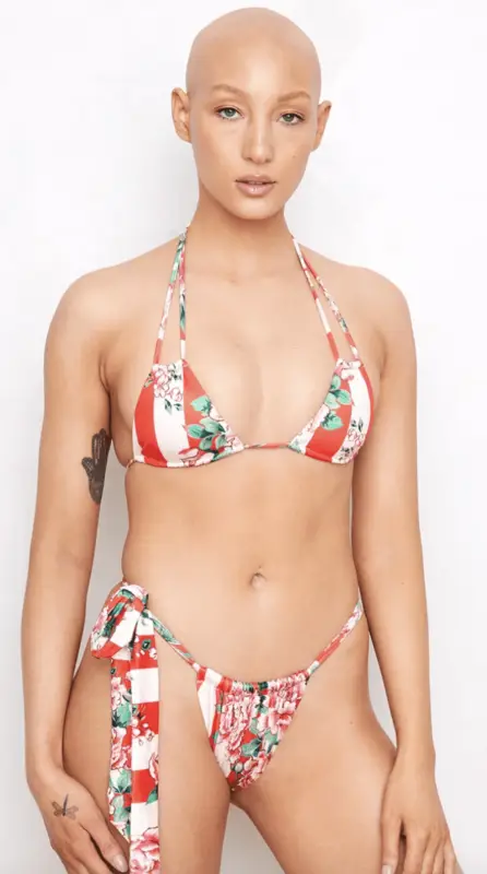 A person with a bald head is wearing a red and white striped bikini with floral patterns, part of the Bikinis That Show You Care About Planet collection. The bikini top has thin straps that tie behind the neck, and the bottom has a side-tie design. They have a tattoo on their left upper arm and are standing against a plain white background.