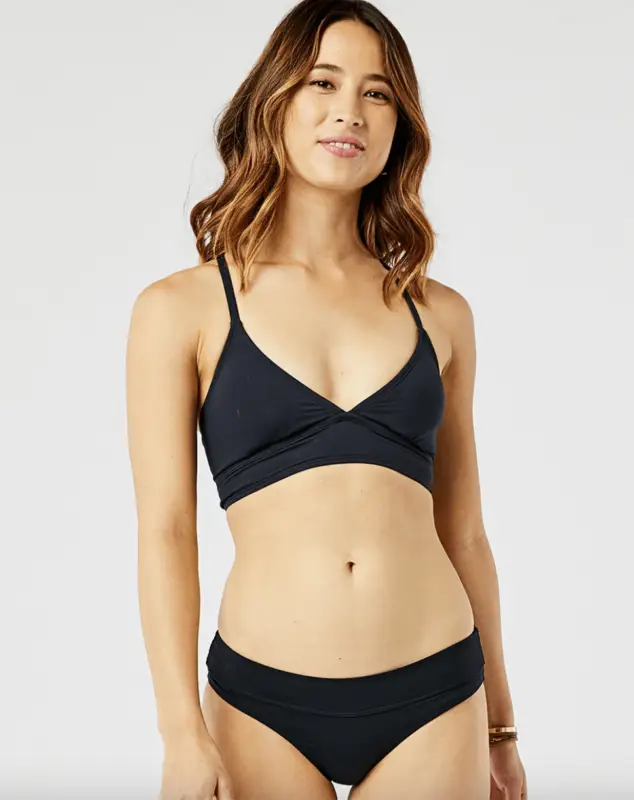 A person stands against a plain background wearing a black bikini from the "Bikinis That Show You Care About Planet" collection. They have shoulder-length, wavy hair and are smiling slightly. Their arms are relaxed by their sides, with one wrist adorned with a bracelet.
