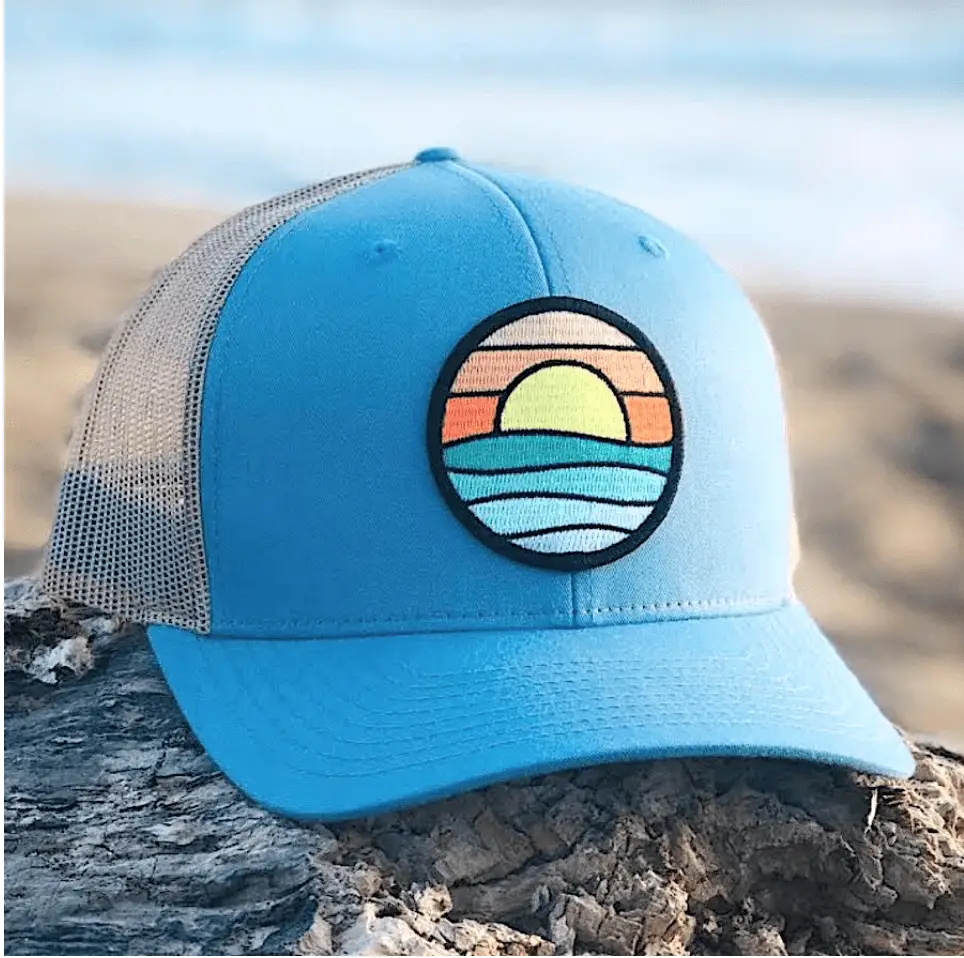 A blue and white mesh trucker hat from Tommy Breeze's apparel collection, featuring an embroidered patch of a colorful sunset over waves, is placed on a weathered log at the beach. The sea and sandy shore are in the blurred background.