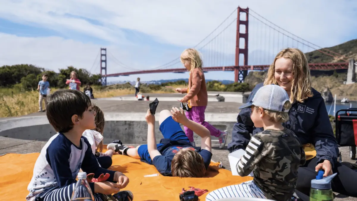 Children and an adult are sitting and playing on an orange blanket outdoors, with the Golden Gate Bridge in the background. The children are engaged in different activities, with one laying down looking at a phone, while the adult is smiling and interacting with them—one of the best things to do in North Bay this March.