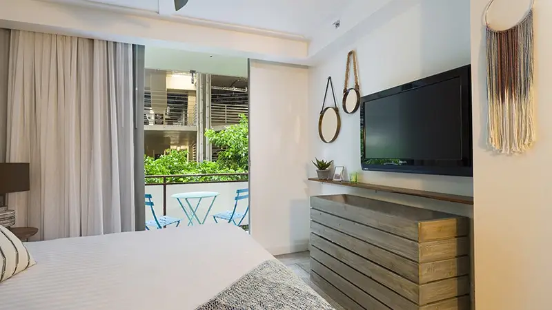 A modern hotel room, among the best hotels on Oahu, features a neatly made bed with white linens, a flat-screen TV mounted on the wall, a wooden dresser, and decorative elements such as hanging mirrors and macramé. The room overlooks a balcony with blue chairs and a view of a lush, green courtyard.
