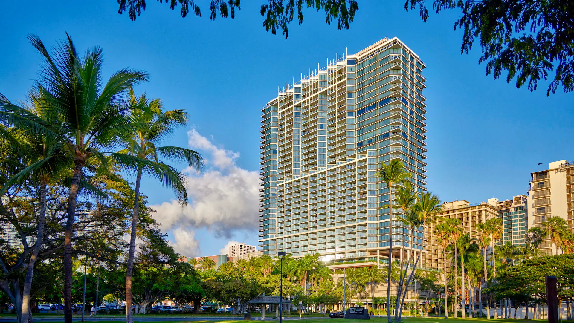 A tall, modern high-rise building stands against a backdrop of a clear blue sky. The building is surrounded by palm trees and lush greenery in the foreground, with other smaller buildings visible in the background. This sunlit scene captures one of the best hotels on Oahu, creating a vibrant atmosphere.