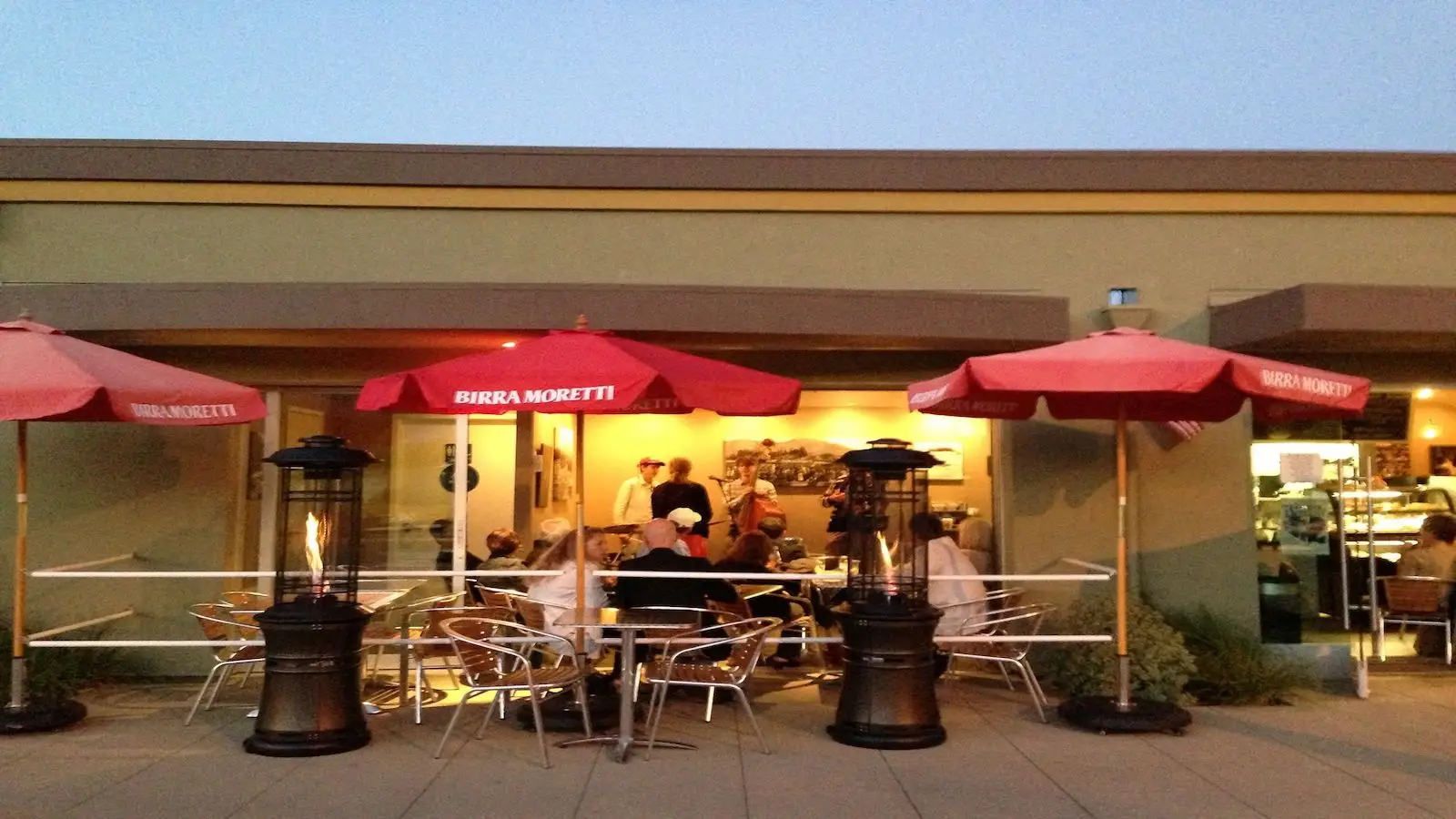 A restaurant patio in the evening light. Several groups of people are seated at metal tables under red umbrellas labeled "Birra Moretti," reminiscent of a scene from King of the Road Bay Area Backroads' Doug McConnell. Heat lamps are positioned near the tables, and patrons inside enhance the cozy atmosphere.