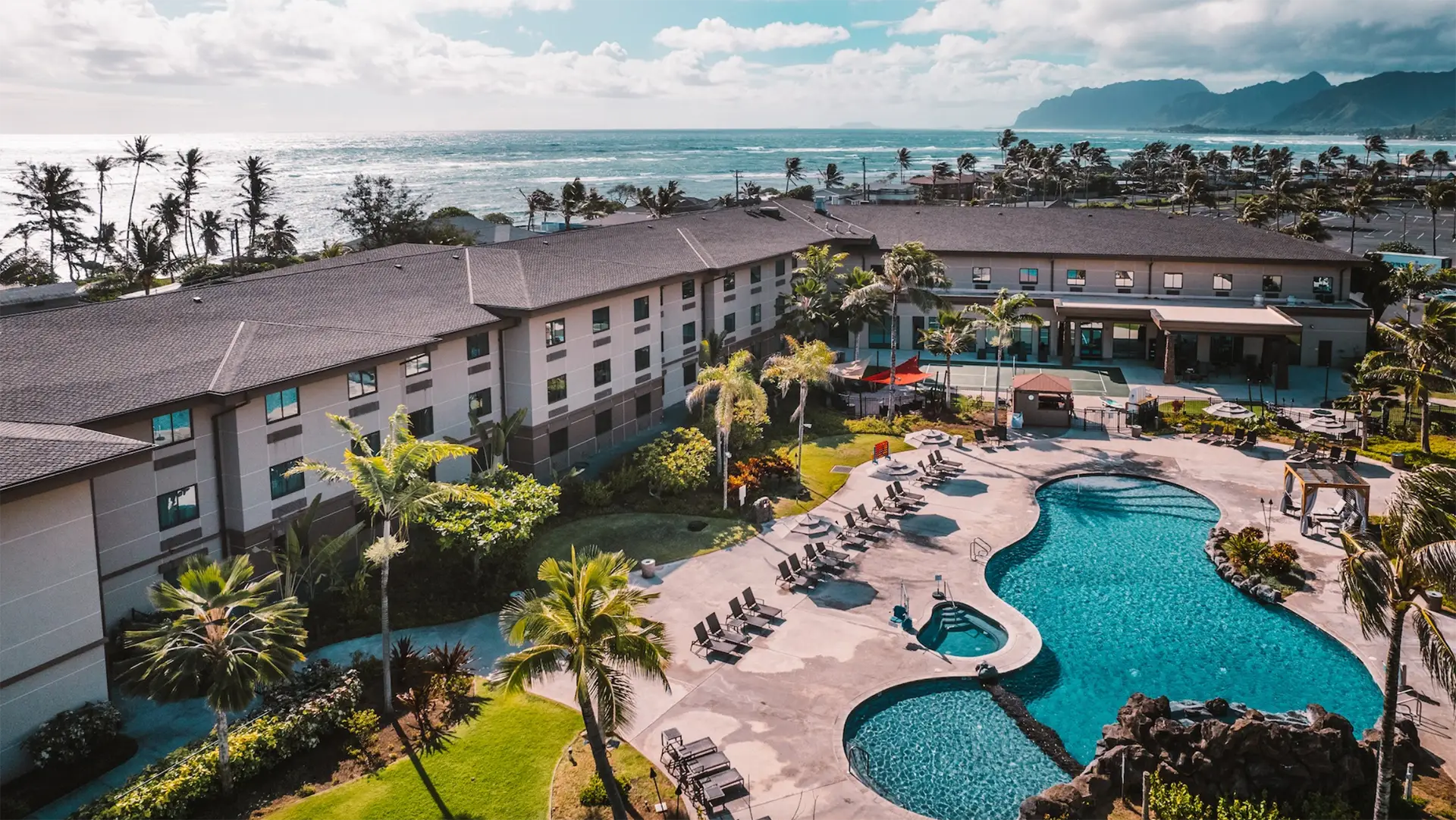 Aerial view of a resort featuring a large, curvy swimming pool surrounded by lounge chairs, palm trees, and landscaped gardens. One of the best hotels on Oahu, the resort is located near the coastline with the ocean and mountains visible in the background under a partly cloudy sky.
