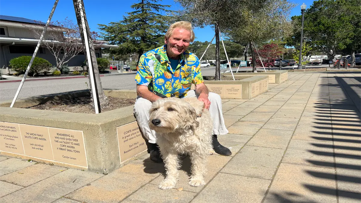 A man wearing a colorful Hawaiian shirt and white pants, reminiscent of King of the Road Bay Area Backroads' Doug McConnell, smiles while crouching beside a fluffy dog. They are outdoors on a sunny day, posing on a paved area with engraved stone benches and abundant greenery in the background.