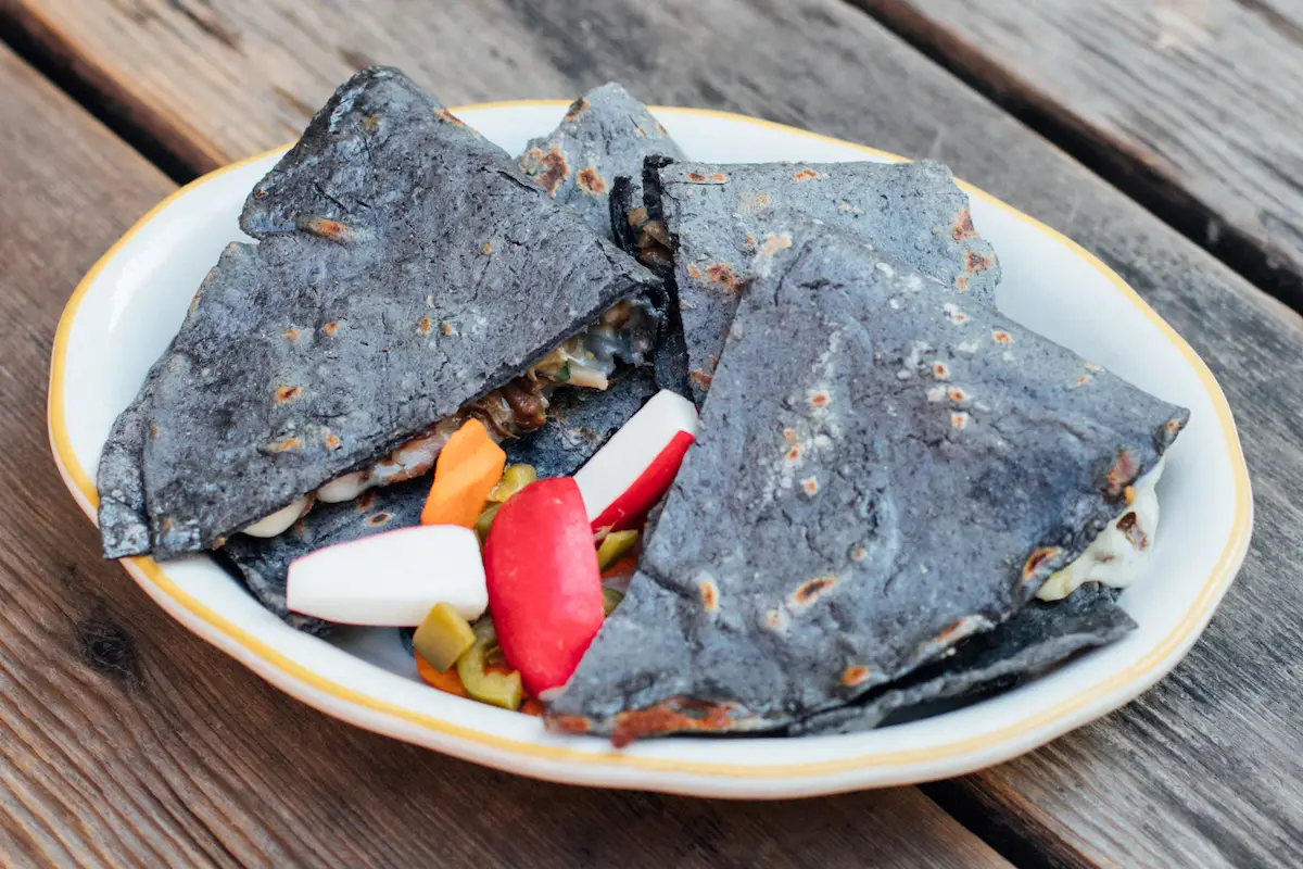A white plate with blue corn quesadillas cut into triangles, served with pickled vegetables including carrots, radishes, and jalapeños. The dish evokes the vibrant flavors often celebrated in Oakland's Michelin restaurants. The plate is on a wooden surface.
