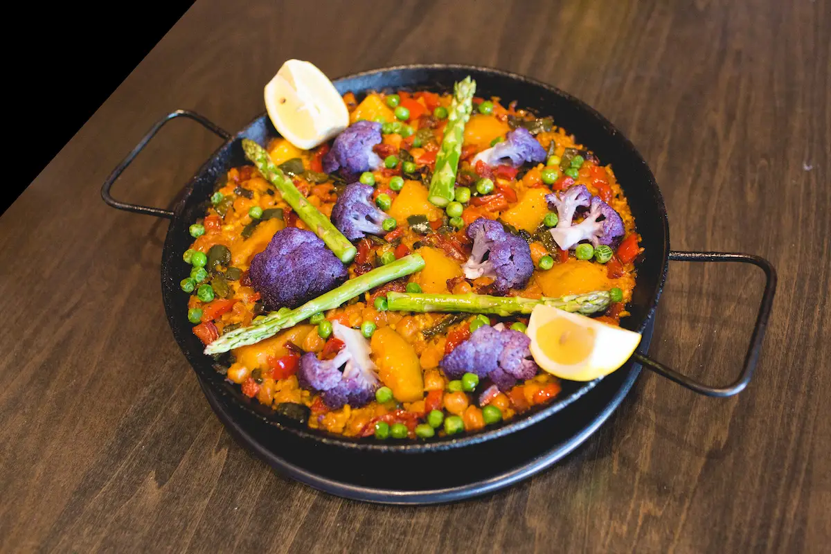 A colorful vegetable paella in a black paella pan on a wooden table, reminiscent of dishes found at Oakland Michelin restaurants. The dish includes vibrant purple cauliflower, yellow bell peppers, green peas, red bell peppers, and asparagus spears, garnished with lemon wedges.