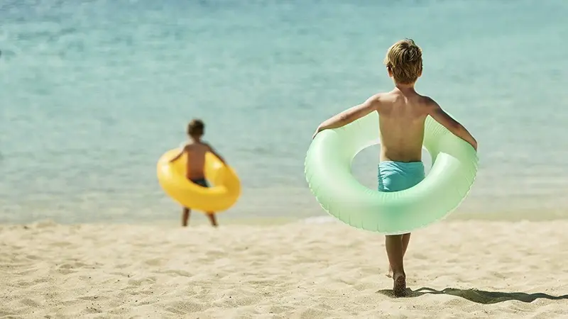 Two children are on a sandy beach by the ocean. The child in the foreground, wearing light blue swim trunks, holds a green inflatable tube around their body. Meanwhile, the child in the background holds a yellow inflatable tube. Both look excited about their day of adventure near some of the best hotels on Oahu.