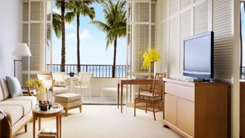 A well-lit living room with wooden furniture and neutral tones. The space opens to a balcony through large white shutters, revealing views of tall palm trees and the ocean, epitomizing a luxury getaway in Hawaii. The room features a TV, a desk, and seating arranged around a small round table.