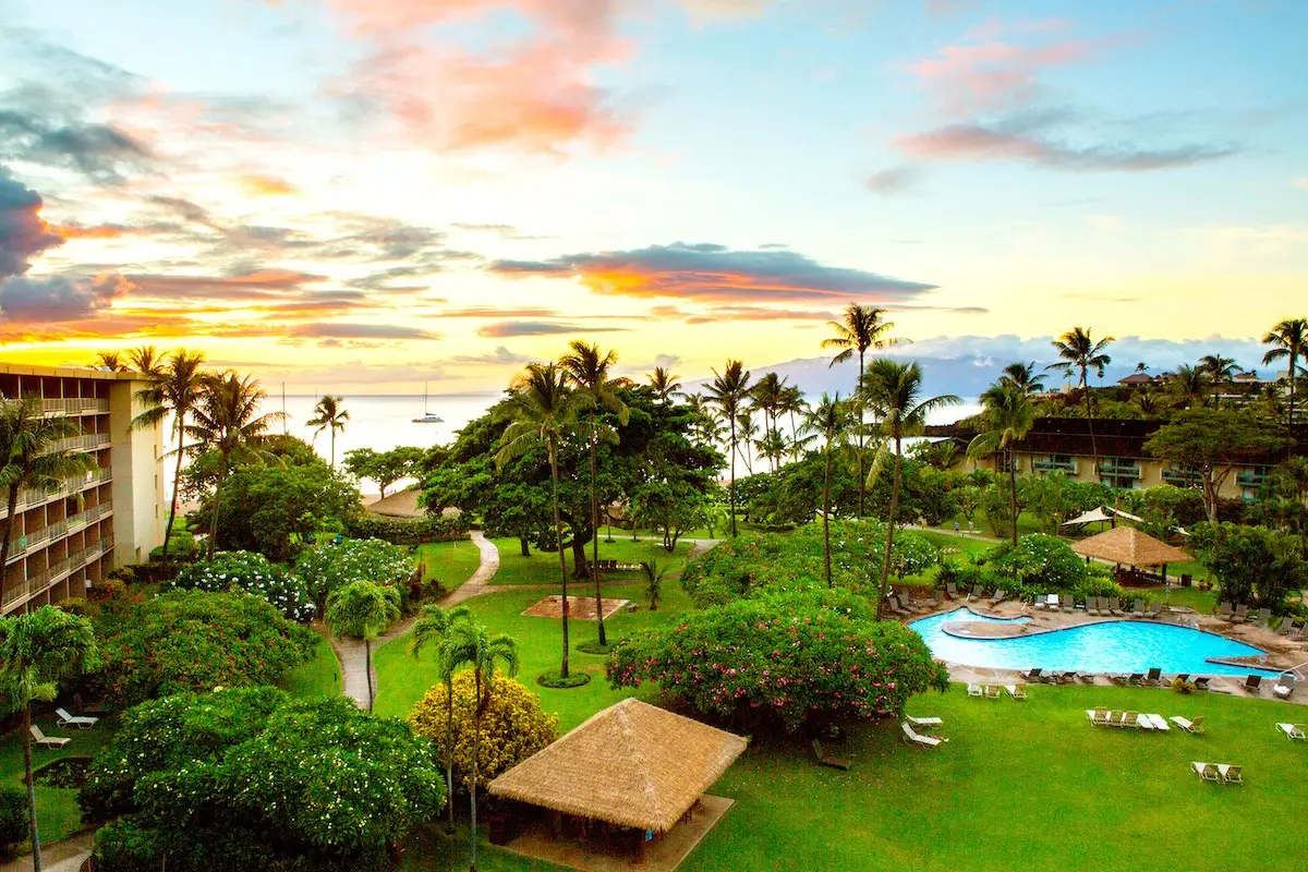 Aerial view of a tropical resort at sunset featuring lush green gardens, palm trees, a winding path, pool, and thatched-roof gazebos. A large building on the left, reminiscent of the Kaanapali Beach Hotel in Hawaii, and the ocean with a distant sailboat under a colorful sky in the background.