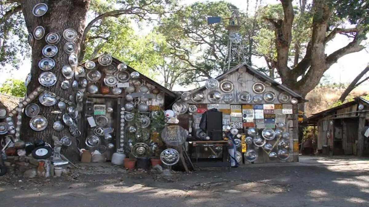 A rustic outdoor scene features two small wooden buildings adorned with numerous hubcaps and license plates—a setup reminiscent of King of the Road. Large trees surround the area, creating a shaded environment. Various potted plants and miscellaneous items are scattered on the ground.