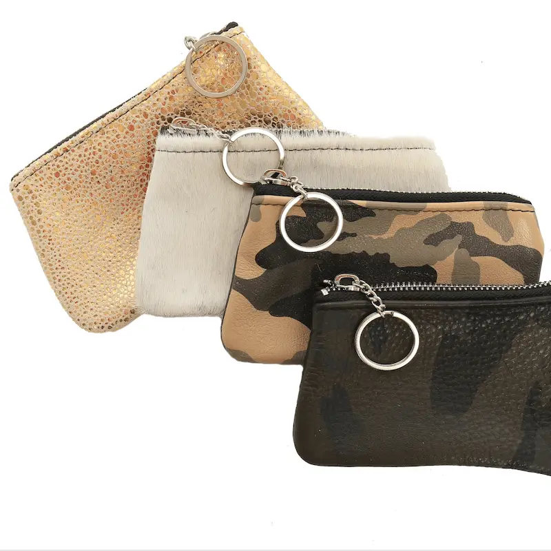 Four small zipper pouches with keyring attachments are shown, designed by Lynn Tallerico. The pouches boast various textures and designs: one is gold and sparkly, one white and furry, one with a camouflage pattern, and one dark and leather-like.
