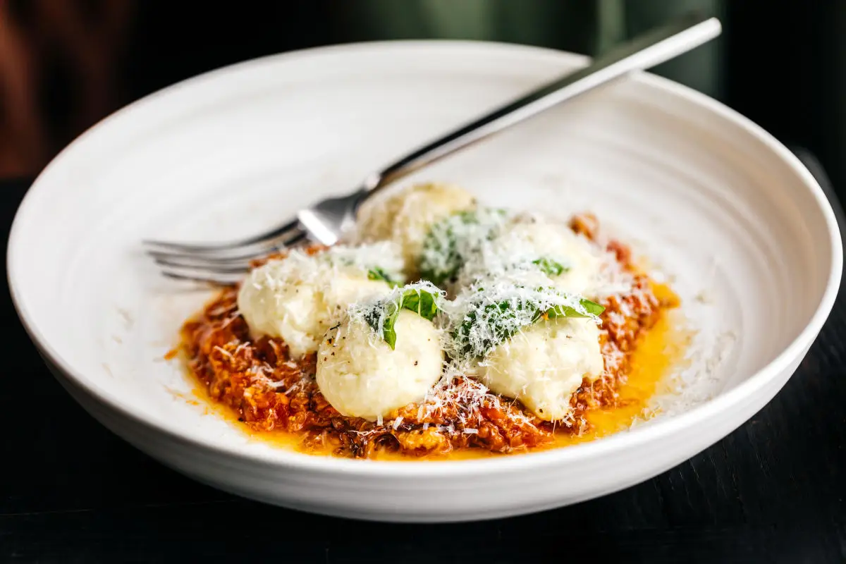 A white plate with a serving of ricotta gnocchi placed on top of a bed of tomato sauce, garnished with grated Parmesan cheese and basil leaves—just like you'd find in Oakland Michelin restaurants. A fork rests on the edge of the plate.