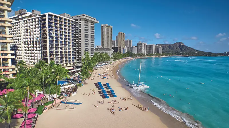 A picturesque beach scene featuring a sandy shore lined with umbrellas and lounge chairs, tall buildings along the coastline, and crystal-clear blue waters. People are enjoying the sun and ocean at this perfect local getaway, with a sailboat docked near the shore and mountains in the background.
