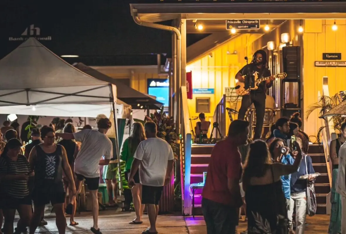 A lively outdoor night market scene in Princeville Kauai with a small crowd of people browsing stalls. A guitarist stands on a brightly lit stage at the back, playing music. The area is illuminated with warm lights, and vendors are set up under tents, offering various items during the annual events.