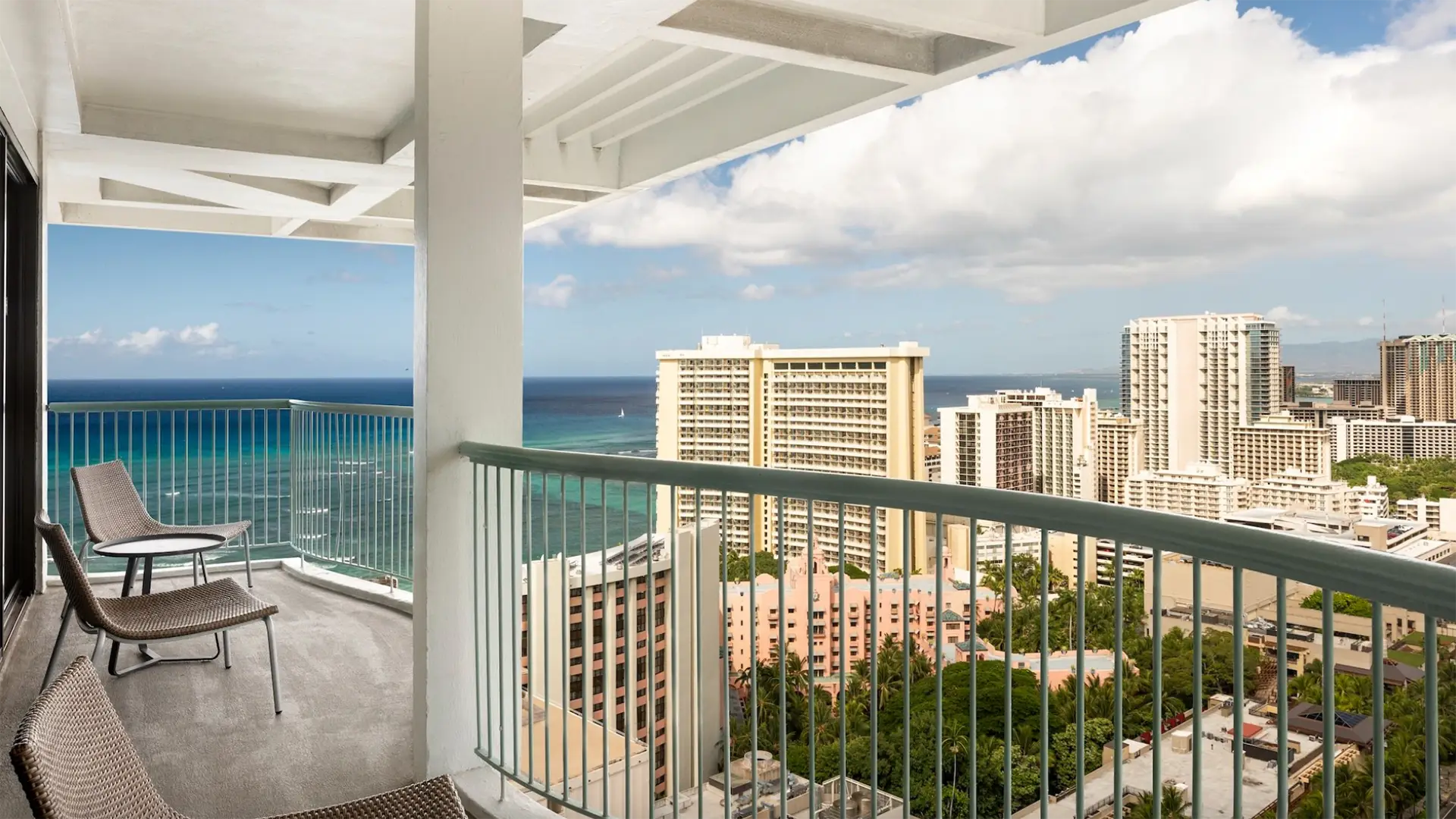 A balcony with two chairs and a small table overlooks a coastal cityscape. Tall buildings stand against the scenic backdrop of a vibrant blue ocean and a partly cloudy sky. Nestled among the best hotels on Oahu, this view captures a perfect blend of urban life and natural beauty.