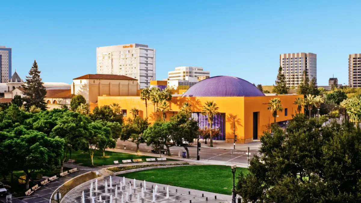 An urban park with a circular water fountain and surrounding trees in the foreground. Behind the park is a bright orange building with a purple dome and palm trees. Several high-rise buildings and a clear blue sky are in the background, making it one of the best things to do in the South Bay this March.
