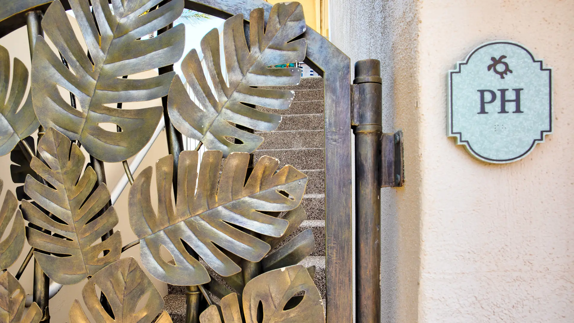 A decorative metal gate with large Monstera leaf designs, evoking a Hawaii home aesthetic, is partially open. Behind the gate, a staircase leads upwards. To the right of the gate, there’s a rectangular sign with the initials "PH," marking The Penthouse Maui.