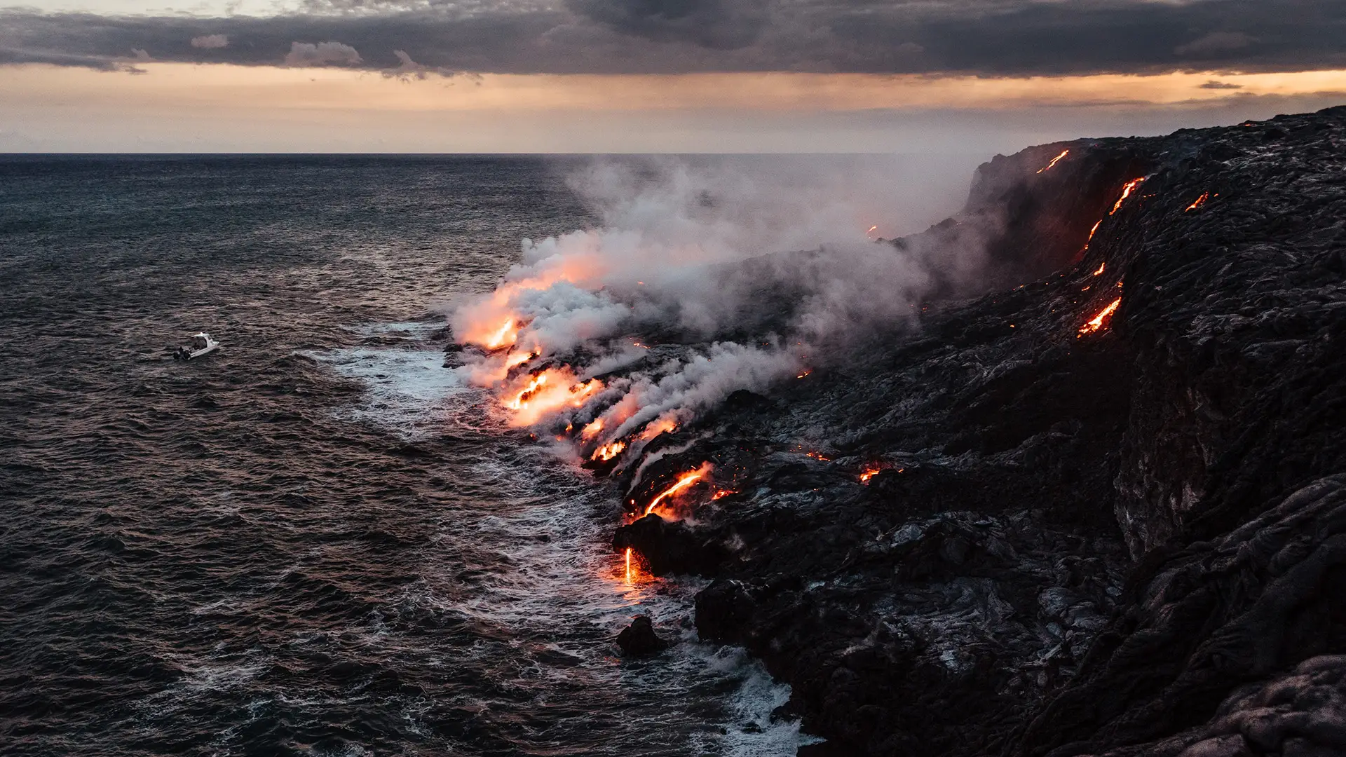A dramatic natural scene showing molten lava flowing into the ocean from a rocky shoreline, creating steam and smoke as they meet. The sky is overcast with a hint of sunset, casting a warm, moody light over the landscape—one of the best places to visit on the Big Island.