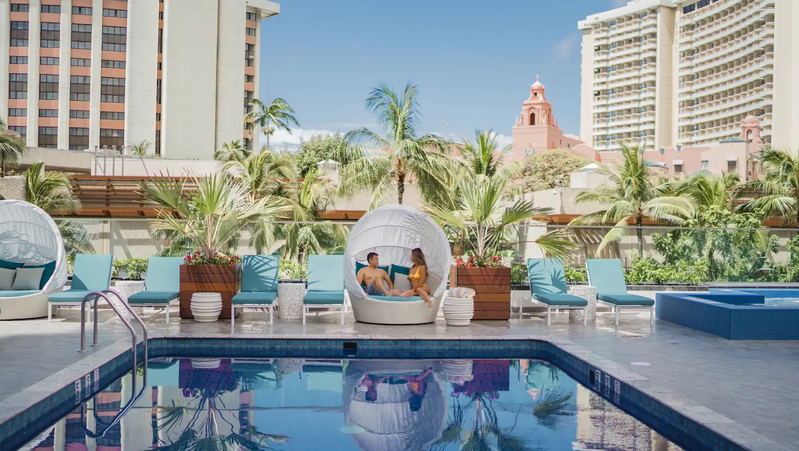 A man and a child relax together in a white pod chair by a pool, surrounded by blue lounge chairs, tropical plants, and tall buildings. The sky is clear and blue, and the pool water reflects the scene. Palm trees and a pink building are visible in the background—one of the best hotels on Oahu.