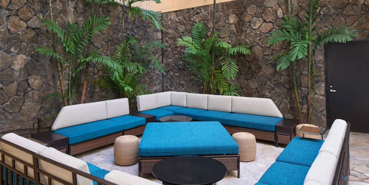 An inviting outdoor seating area with a stone wall backdrop features an L-shaped arrangement of blue and white cushioned sofas. Two small beige poufs, a square blue ottoman, and a round black coffee table are positioned in the center. Lush green plants add a touch of nature.