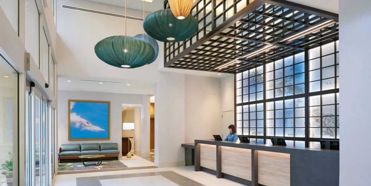 A modern hotel lobby with high ceilings and geometric light fixtures. A reception desk with a staff member is on the right, while the left side has a seating area with a painting on the wall. The floor features a combination of light and dark tiles.