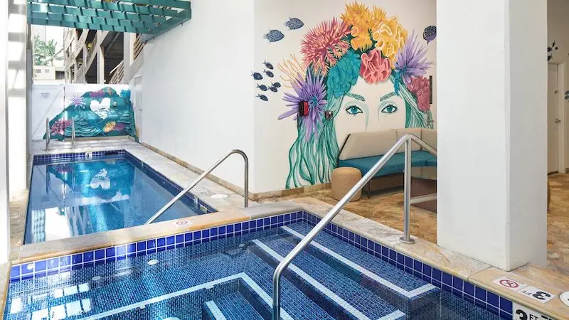 A vibrant indoor pool area at one of the best hotels on Oahu features a hot tub with blue-tiled steps. The wall behind it displays a colorful mural of a woman's face adorned with flowers and sea creatures. A wicker lounge chair and a door leading to another room can be seen in the background.