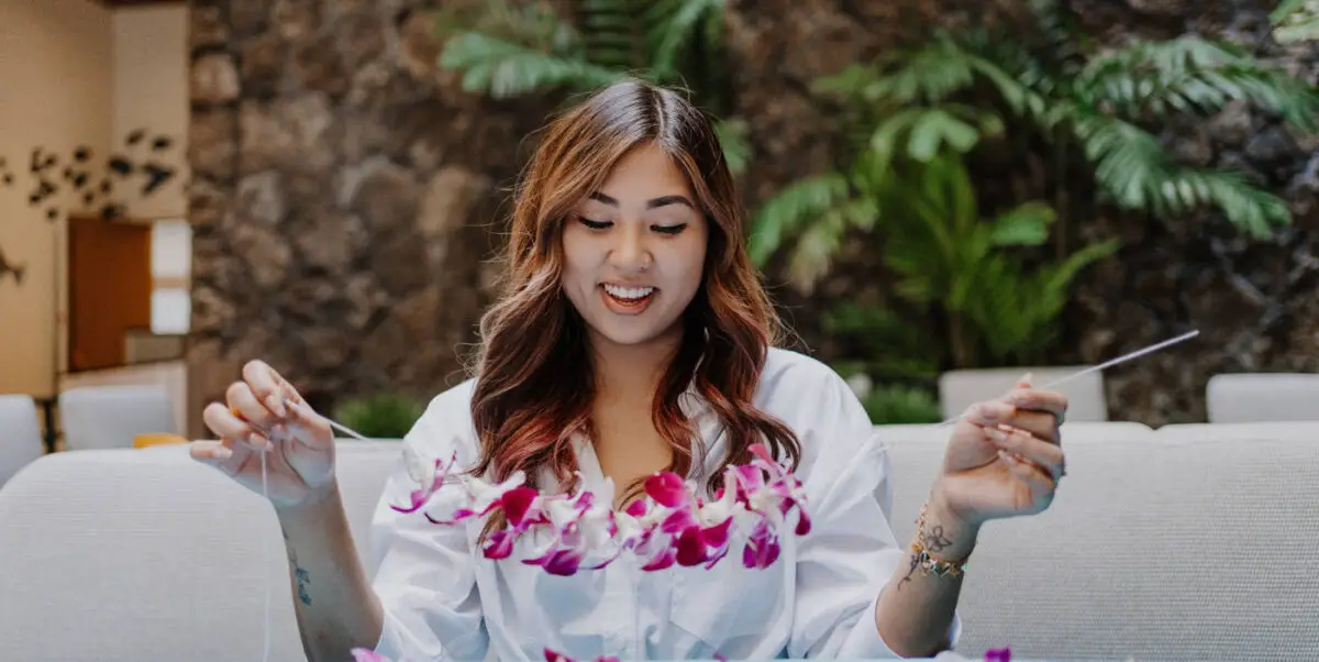 A young woman with long brown hair and a white shirt smiles while threading a pink and white flower lei. She is seated on a light-colored couch in a room adorned with greenery and stone walls in the background.