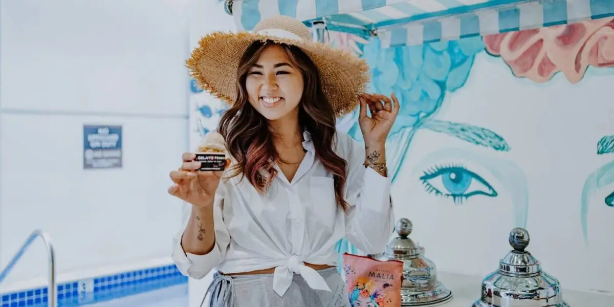 A woman smiles while holding a small cup of gelato. She is wearing a wide-brimmed straw hat and a tied white blouse. Behind her is a vibrant mural and an ice cream stand with silver dispensers and a pink product package.