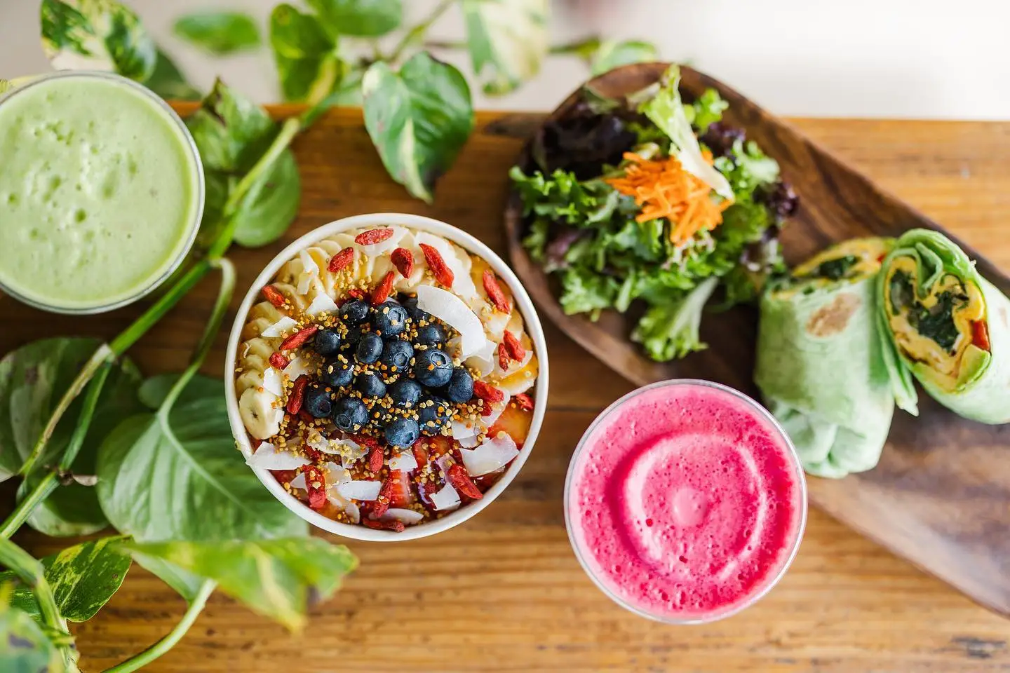 A vibrant meal spread features a smoothie bowl topped with blueberries, nuts, and seeds, alongside a green smoothie, a red smoothie, a small wooden dish with a green salad, and a wrap filled with vegetables on a wooden surface surrounded by green leaves—truly the best lunch North Shore has to offer.