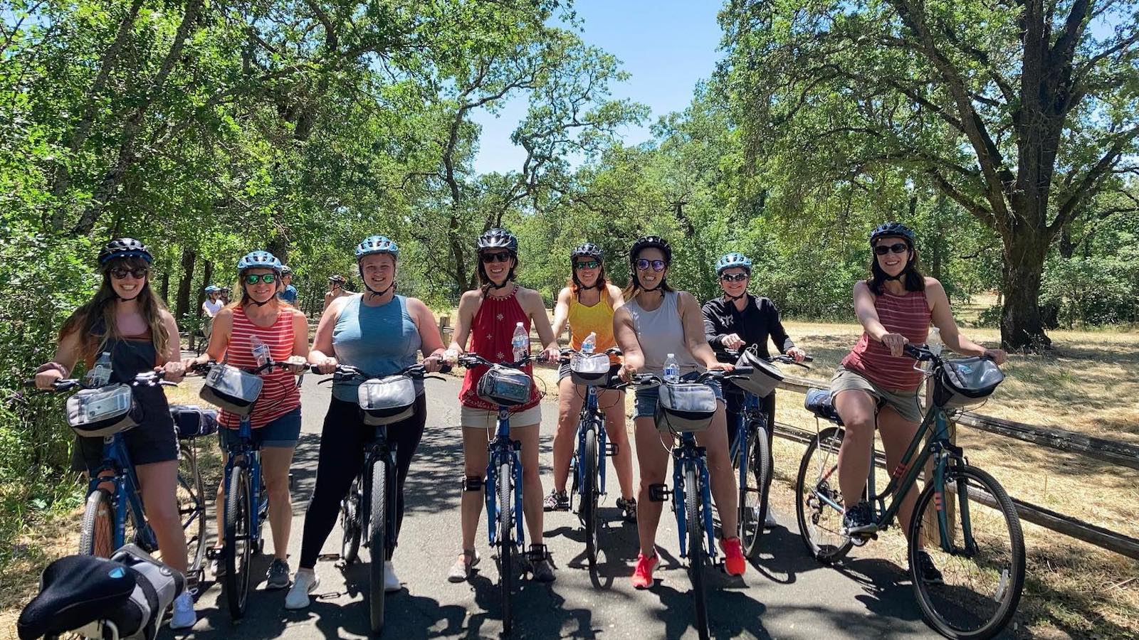 A group of eight people wearing helmets, sunglasses, and casual clothes are riding bicycles on a paved path through a forested area. The weather is sunny, and the group appears to be enjoying one of the top activities in Sonoma.