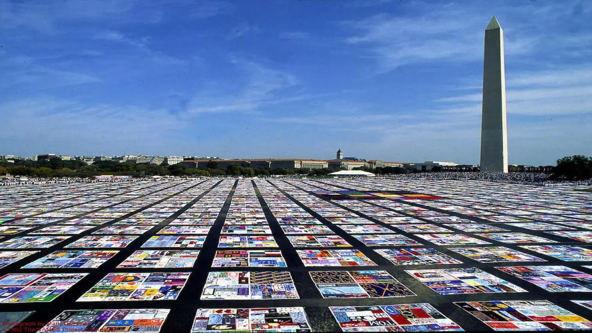 The image shows the Washington Monument with a clear blue sky in the background. In the foreground, a vast field is covered with colorful quilt panels from the NAMES Project AIDS Memorial Quilt, celebrating San Francisco Pride all month long with these special events.