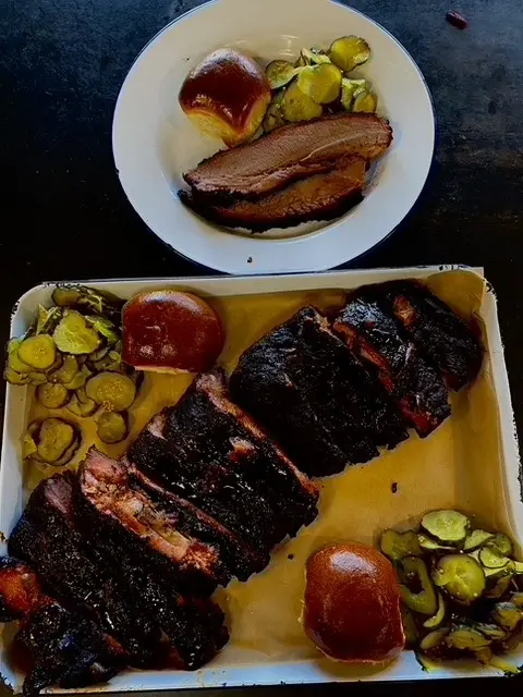 A tray of dark, charred barbecued meat next to pickles and two buns evokes the spirit of a Memorial Day BBQ. Above the tray, a white bowl contains a thick slice of meat and more pickles, with barbecue sauce drizzled on top.